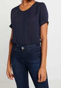 Navy blue blouse with short sleeves, featuring a round neckline and delicate lace trim at the sleeves. Paired with dark denim jeans.