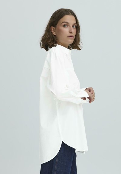 Balta marškiniai su ilgomis rankovėmis, išlenktu apatine dalimi ir standžiu apykakle. Lygus audinys ir laisvas siluetas, derinamas su tamsiais džinsais.