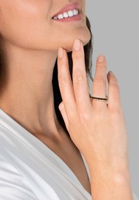 Bague en or avec une rangée de pierres précieuses sombres, portée sur une main reposant sur la mâchoire. Peau lisse et vêtement de couleur claire en arrière-plan.