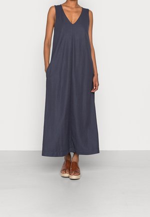Robe maxi bleu marine sans manches avec un profond décolleté en V et des poches latérales, portée avec des sandales compensées en cuir tressé marron sur un fond uni.