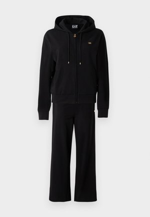 Zwarte rits-hoodie met drawstrings en bijpassende wijde broek, met kleine gouden logo-details op de borst en heup.