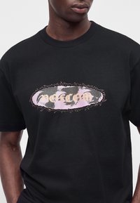 Chemise noire à manches courtes portée par une personne, avec un graphisme ovale aux formes abstraites et le mot "Volcom" en couleurs claires sur la poitrine.