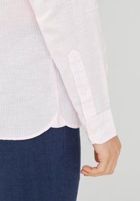 Lichtroze gestreepte shirt gemaakt van lichtgewicht stof, met knoopmanchetten en een gebogen zoom. G gedragen met donkerblauwe broeken.