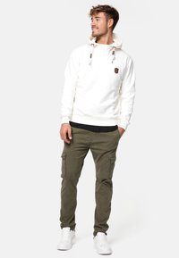 Sudadera blanca con capucha y bolsillo canguro, combinada con pantalones cargo oliva y zapatillas deportivas blancas. El modelo está de pie con las manos en los bolsillos.