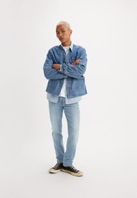 Tânăr cu părul vopsit blond stă cu brațele încrucișate, purtând o jachetă din denim stratificată, blugi de culoare albastru deschis, cămașă albă și adidași negri.