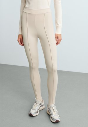 Modèle portant un legging crème taille haute et un haut assorti à manches longues, avec des baskets blanches et grises.