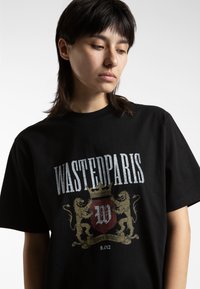 Černé bavlněné tričko s grafickým designem, na kterém je nápis "WASTEDPARIS" v opotřebované typografii, motivy koruny a lva s barevnými akcenty zlata a červené.