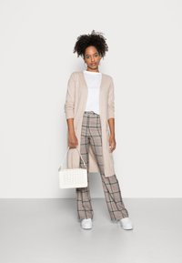 Langer beiger Cardigan, weißes T-Shirt, karierte weit geschnittene Hose in Grau und Beige, hält eine weiße strukturierte Handtasche, kombiniert mit weißen Turnschuhen.