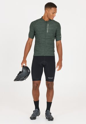 Short-sleeve dark green cycling jersey with a zip front, paired with black padded shorts, black cycling shoes, and a black helmet in hand.