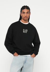 Svart hettegenser med «GAP»-logo i hvitt, ribbestrikket rund hals, avslappet passform og lange ermer, kombinert med lyseblå jeans. Enfarget bakgrunn.