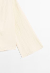 Tissu beige clair texturé avec de larges manches fluides ; forme rectangulaire avec un ourlet brut. Design minimal, apparence douce.