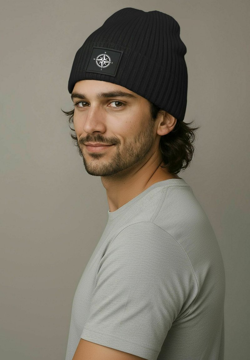 Black ribbed beanie with a flat, circular patch featuring a compass design. Worn with a light gray, short-sleeve shirt against a neutral background.