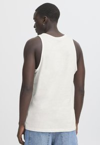 Gaiši bēšs krekliņš bez piedurknēm ar apaļu kakla izgriezumu, brīvs piegriezums un mīksts kokvilnas materiāls. Aizmugures skats parāda vienkāršu dizainu bez redzamām detaļām.