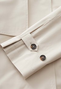 Camicia beige chiaro con colletto a punta, polsini con bottoni scuri e dettagli cuciti sulle spalle. Tessuto liscio.