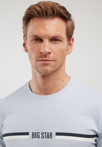 Gaiši zils kokvilnas T-krekls ar apaļu kakla izgriezumu, īsām piedurknēm un horizontalām svītrām, uz krūtīm ir teksts "BIG STAR".