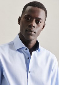 Ung sort mand med kort hår iført en lyseblå skjorte med knapper, der roligt ser mod kameraet foran en ensfarvet baggrund.