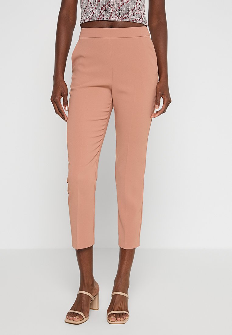 Spodnie w kolorze brzoskwiniowym, dopasowane, o gładkiej fakturze, w wąskim kroju i skróconej długości, zestawione z wzorzystą bluzką bez rękawów oraz jasnymi butami.