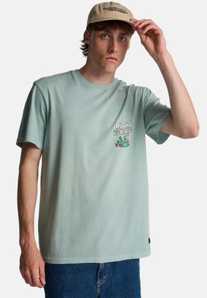 Młody mężczyzna w jasnozielonej koszulce z nadrukiem i beżowej czapce, poprawiający daszek prawą ręką, na jednolitym białym tle.