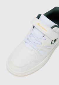 Baskets blanches avec une tige en cuir lisse, des lacets bleu clair, une doublure en tissu vert et un logo "Champion" doré sur la sangle.