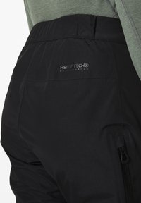 Czarne spodnie robocze na zewnątrz z elastycznym pasem, zamykaną na zamek kieszenią boczną oraz srebrnym brandingiem "Helly Tech Performance" nad kieszenią.