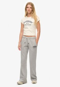 Pantalons de survêtement gris clair avec cordon de serrage et poches, associés à un t-shirt blanc court avec un logo et un texte "SUPERDRY" bleu marine.