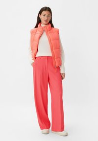 Coral puffer vest over a cream sweater, paired with bright coral wide-leg trousers. Smooth texture, casual design, zipper details, and white sneakers.
