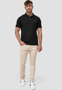 Sort poloskjorte med knapper og lille rødt logo, kombineret med lyse beige slim-fit bukser og hvide sneakers med sorte accenter.