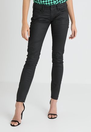 Mujer vestida con pantalones negros ajustados y sandalias de tacón negro, de pie contra un fondo claro y sencillo.