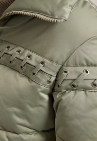 Veste matelassée vert clair avec fermeture éclair, lacets décoratifs avec œillets en métal et texture lisse sur le tissu.