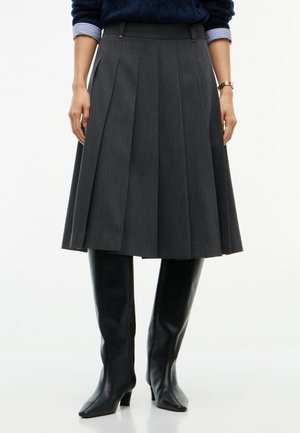 Femme portant une jupe plissée sombre à hauteur du genou, des bottes noires hautes jusqu'au genou, un pull bleu marine et une chemise rayée en dessous.