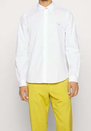 Homme portant une chemise blanche à manches longues avec un petit logo brodé et un pantalon jaune vif sur un fond uni.