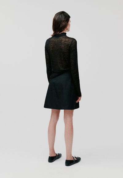 Haut noir à manches longues avec une texture transparente, associé à une jupe noire. Le mannequin porte des ballerines noires ; le fond est neutre et minimal.