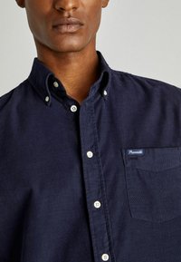 Camisa de algodón azul marino de botones con un bolsillo en el pecho, botones blancos y etiqueta de marca en el bolsillo, con un acabado de tejido texturizado.