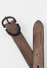 Bruine leren riem met een matte zwarte metalen gesp. Voorzien van lichte stiksels en meerdere gaatjes voor maatverstelling. Merkennaam in reliëf.