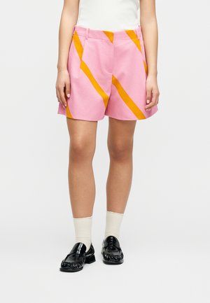 Άτομο που φοράει ροζ σορτς με πορτοκαλί διαγώνιες ρίγες, λευκές κάλτσες και μαύρα μοκασίνια, στέκεται μπροστά σε ένα απλό λευκό φόντο.