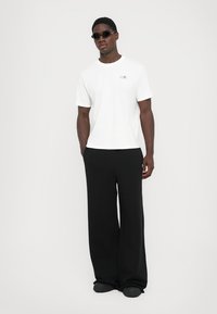 T-shirt de algodão branco com logotipo, combinado com calças largas pretas e sapatos slip-on pretos. Modelo usa óculos de sol pretos.