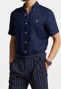 Navy blue short-sleeve button-down shirt in lightweight linen with a logo on the chest, paired with pinstriped navy trousers.