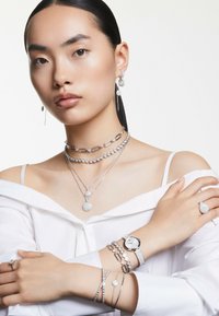 Silver smycken som visas inkluderar lager på lager-halsband, armband och örhängen med intrikata mönster. En silverklocka kompletterar ensemble.