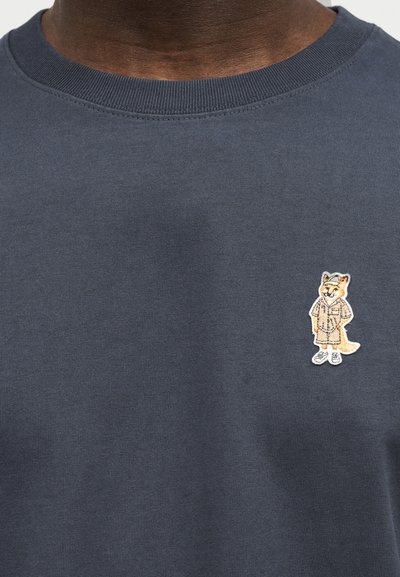 Σκούρα γκρι μπλούζα με στρογγυλή λαιμόκοψη που φέρει ένα μικρό κεντημένο χαρακτήρα αλεπούς με καπέλο, σακάκι, σορτς και αθλητικά παπούτσια στο αριστερό μέρος του στήθους.