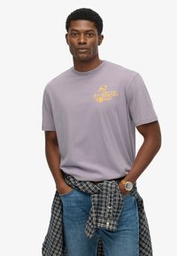 Camiseta de algodón color púrpura claro con un logo amarillo en el pecho, combinada con jeans azules y una camisa de cuadros atada a la cintura.