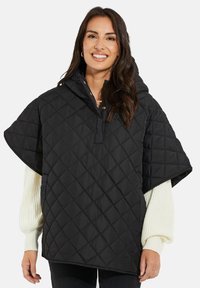 Svart vadderad poncho med huva, med en fjärdedels knappstängning och breda ärmar, lagd över en krämfärgad tröja.