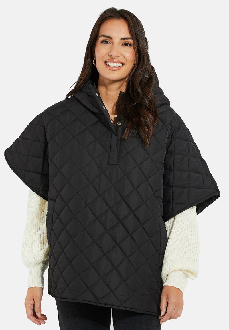 Svart vadderad poncho med huva, med en fjärdedels knappstängning och breda ärmar, lagd över en krämfärgad tröja.