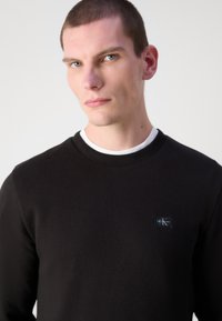 Svart strikket sweatshirt med rund hals, teksturert stoff og en liten logo-patch på venstre bryst. Slim-fit design med et komfortabelt utseende.