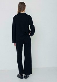 Top negro de manga larga con un corte relajado, combinado con pantalones anchos negros y zapatos planos negros, presentado desde atrás sobre un fondo claro.