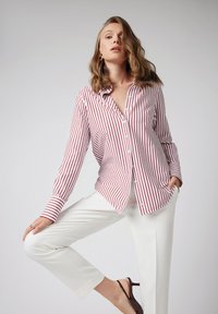 Kobieta w czerwono-białą pasiastej koszuli z guzikami, białych spodniach i ciemnobrązowych szpilkach, pozująca z jedną ręką w kieszeni na tle jednolitego tła.