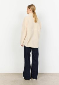 Beige fleece-takki, jossa on korkea kaulus, löysä malli ja teksturoitu pinta, yhdistetty tummansinisiin kellotaskuhousuihin ja valkoisiin lenkkareihin.