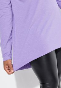 Helllila Langarmoberteil mit abgerundetem Saum und leicht strukturiertem Stoff. Kombiniert mit eng anliegenden schwarzen Leggings, die eine glatte Optik zeigen.