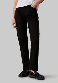 Pantalones negros con un diseño de pierna recta, hechos de mezclilla, que cuentan con un bolsillo frontal y una textura sutil. Combinados con zapatos negros brillantes.