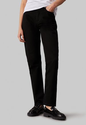 Schwarze Hose mit geradem Bein, aus Denim, ausgestattet mit einer Fronttasche und einer subtilen Textur. Kombination mit glänzenden schwarzen Schuhen.