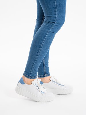 Persona con vaqueros azules y zapatillas blancas con detalles azules, de pie con las piernas cruzadas sobre un fondo blanco.